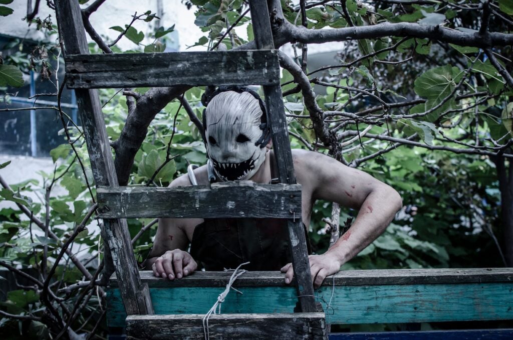 A spooky masked figure peers through a ladder in an eerie overgrown garden setting.